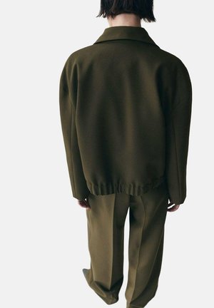 Giacca verde scuro oversize con colletto ed elastico al fondo, abbinata a pantaloni coordinati a gamba larga, realizzati in tessuto liscio.