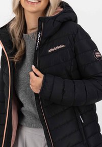Schwarze Steppjacke mit Kapuze, mit pinken Akzenten am Reißverschluss und im Innenfutter, sowie Markenlogos auf der Brust und dem Ärmel.