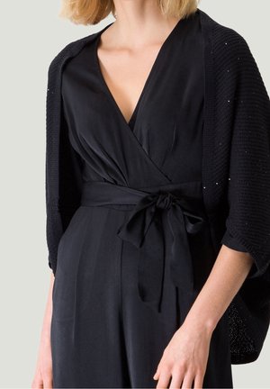 Femme portant une robe portefeuille noire avec un nœud à la taille et un châle en tricot noir sur les épaules, cadrée au-dessus des épaules.