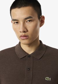 Läheltä kuvattuna nuori mies, jolla on lyhyet mustat hiukset, pukeutuneena ruskeaan poolopaitaan, jossa on kauluksessa pieni vihreä krokotiilin logo.