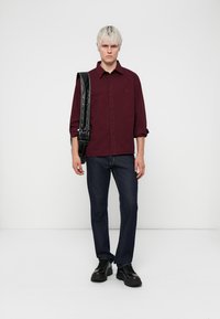 Bordeaux skjorte med knapper, mørkeblå jeans, sorte sko og en sort naglet skuldertaske. Model står foran en ensfarvet hvid baggrund.
