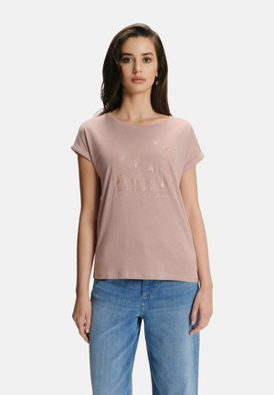 Kobieta z długimi ciemnymi włosami, ubrana w jasnoróżową koszulkę z krótkim rękawem z kwiatowym wzorem oraz jasnoniebieskie dżinsy, stojąca na jednolitym tle.