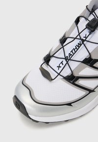 Białe sportowe buty z srebrnymi akcentami, teksturowana siateczka na wierzchu i czarny system sznurowania. Posiadają wyraźną, zakrzywioną podeszwę dla lepszej przyczepności.