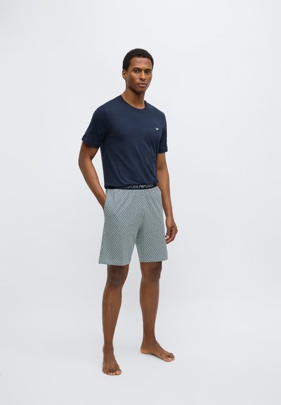 Námornícka bavlnená tričko s krátkymi rukávmi; vzorované šortky s geometrickými prvkami a elastickým pásom. Model stojí bosý na svetlom pozadí.