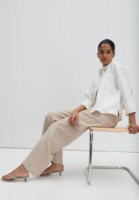 Donna seduta di lato su una sedia, indossando una camicia bianca, pantaloni beige a gamba larga e sandali con tacco basso e punta aperta, su uno sfondo chiaro.