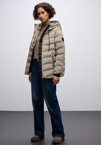 Beige dunjakke med hætte, sorte accenter og lynlåse i siderne, båret over en brun sweater og parret med blå jeans og brune støvler.