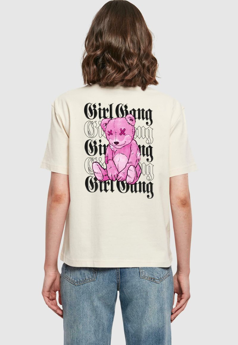 Kremo spalvos per didelės t-shirt su rožiniu animaciniu lokiu ir ryškiu juodu tekstu "Girl Gang" sudėtu ant nugaros.