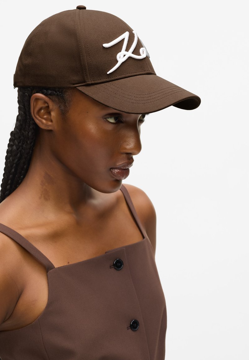 Γυναίκα με πλεξούδες φοράει καφέ καπέλο με λευκή γραφή και καφέ αμάνικο τοπ με μαύρα κουμπιά, κοιτάζοντας δεξιά σε λευκό φόντο.