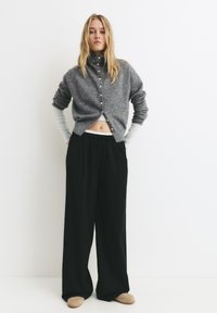 Sivý pletený cardigan s vysokým golierom a detailmi na gombíky, kombinovaný s čiernymi nohavicami s širokými nohavicami. Oblečenie má vrstvený vzhľad.