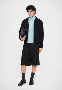 Marineblå lynlåsjakke over en lyseblå rullekrave, kombineret med sorte shorts og sorte sko. Holder en tekstureret sort taske.