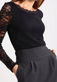 Czarny koronkowy top z długimi rękawami, kwiatowym wzorem i falistym dekoltem, zestawiony z wysokim stanem szarymi spodniami z plisami.