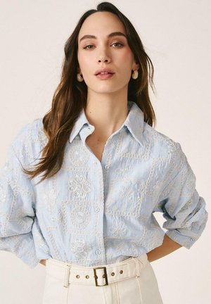 Joven con cabello largo y castaño, vistiendo una camisa de botones azul claro con bordados y pantalones blancos con cinturón, posando con las manos en las caderas.