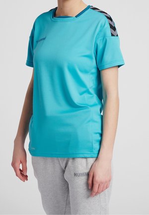 Persona que lleva una camiseta deportiva azul brillante de manga corta con rayas Chevron negras en los hombros y pantalones deportivos grises con el logotipo de la marca.