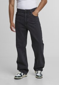 Bărbat purtând blugi negri largi, maiou alb și adidași verzi cu alb, stând cu o mână în buzunar.