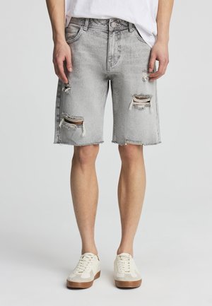 Persona che indossa pantaloncini di jeans strappati grigio chiaro, sneakers bianche e una maglietta bianca, in piedi davanti a uno sfondo grigio chiaro uniforme.