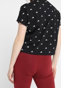 Černý top s krátkými rukávy z látky s bílým trojúhelníkovým vzorem. Má zaoblený spodní lem a detail v podobě zadního dílu.