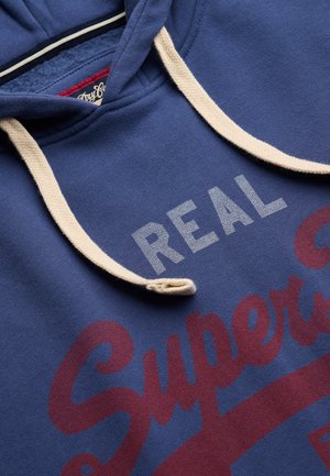 Blå hættetrøje med creme hættekordel med teksten "REAL Superdry" i hvide og røde bogstaver på fronten.