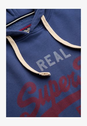 Blå hættetrøje med creme hættekordel med teksten "REAL Superdry" i hvide og røde bogstaver på fronten.