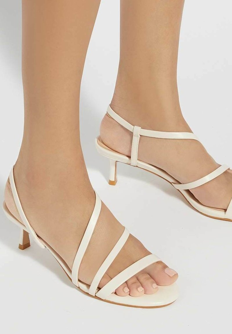 Sandales à talons hauts beiges avec plusieurs fines lanières, design à bout ouvert et petit talon carré. Texture lisse avec une esthétique minimaliste.