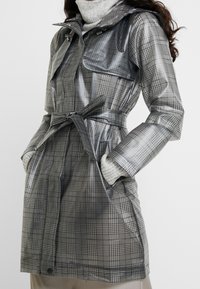 Šedý károvaný nepromokavý trenčkot s páskem v pase, vysokým límcem a dvěma bočními kapsami. Vyznačuje se lesklou, hladkou texturou a nastavitelnými manžetami.