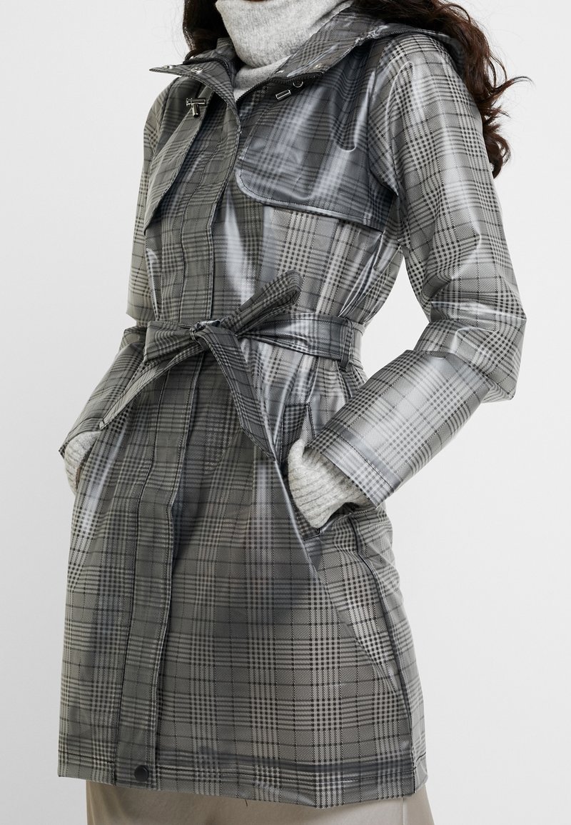 Šedý károvaný nepromokavý trenčkot s páskem v pase, vysokým límcem a dvěma bočními kapsami. Vyznačuje se lesklou, hladkou texturou a nastavitelnými manžetami.