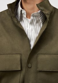 Giacca in suede verde oliva con colletto alto, due tasche frontali e chiusure a pressione, indossata sopra una camicia bianca con righe grigie.