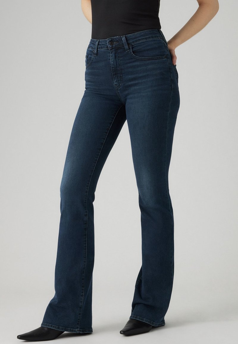 Femme portant un jean bootcut bleu foncé taille haute et des chaussures noires à bout pointu, debout devant un fond gris uni.