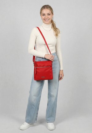 Young woman wearing white turtleneck and jeans stands smiling, holding a red crossbody bag with adjustable strap and front zipper pocket.