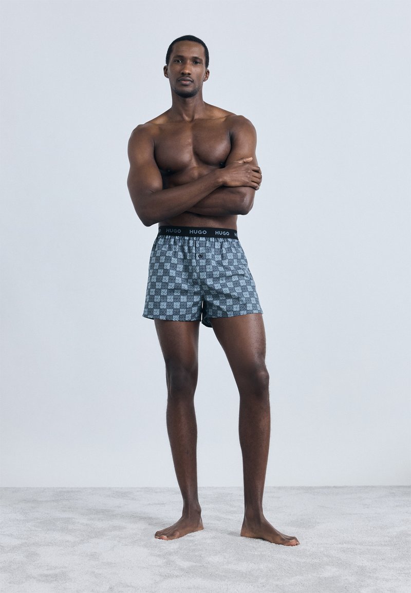 Μπόξερ σορτς από ύφασμα με γκρι σχέδιο, με μαύρη ελαστική μέση που φέρει το λογότυπο "HUGO" και χαλαρή εφαρμογή. Το μοντέλο ποζάρει ξυπόλητο σε γκρι επιφάνεια.