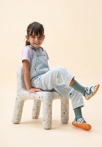 Kindersessel aus strukturiertem, gesprenkeltem Recyclingmaterial. Er hat abgerundete Kanten und stabile Beine. Das Kind trägt eine helle Latzhose und Sneakers.
