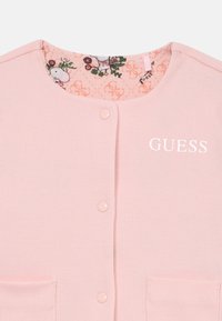 Ružičasti rebrasti kardigan s četiri dugmeta i dva džepa. Unutrašnja podstava sadrži cvjetni uzorak i uzorak s logotipom. "GUESS" logotip otisnut u bijeloj boji.