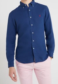 Mörkblå långärmad skjorta med knappar och krage, med vita knappar och en liten röd logotyp på vänster bröst. Slät textur.