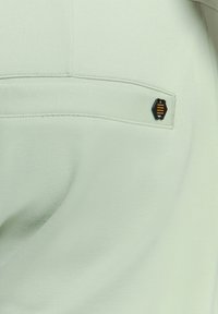 Lichtgroene stof met een gladde textuur, voorzien van een zwart hexagonaal logo met gele accenten op een achterzak.