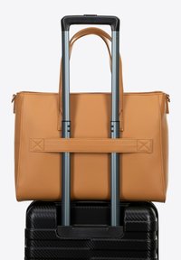 Bolso tote de cuero color marrón claro con dos asas, colocado sobre una maleta negra texturizada con asa telescópica. Bolsa asegurada por una correa de tela.