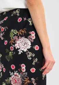 Svart blommig kjol med en mängd rosa och gröna blommor, med intrikata detaljer och en slät textur. Synlig hand nära fållen.