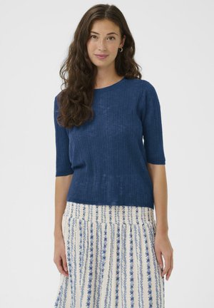 Femme aux cheveux longs et ondulés portant un haut bleu à manches courtes côtelé et une jupe blanche à rayures florales verticales bleues, debout devant un fond uni.