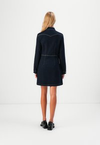 Blond kvinne som står vendt bort, iført en mørk denimkjole med lange ermer og svarte hæler mot en enkel hvit bakgrunn.