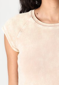 Light pink fitted t-shirt with a round neckline and cap sleeves. Features a textured, slightly faded fabric and contrasting stitching details.