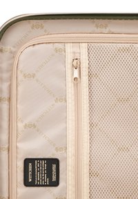 Interno di un bagaglio da viaggio beige con una tasca con cerniera, un compartimento in rete e un motivo a logo dorato. Presenta un'etichetta di cura nera. Tessuto texturizzato.