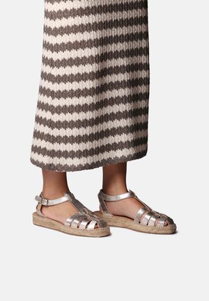 Noge v srebrnih kovinskih sandalih z obliko črke T in tkanimi podplati espadrij, kombinirane s krilom srednje dolžine v rjavo-bež črtah.