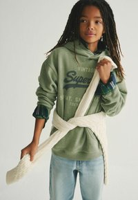 Sudadera verde con el logo de "Superdry" y dobladillo de canalé, combinada con jeans de mezclilla claros. Envueltos en una bufanda de punto en color crema. Cabello oscuro y texturizado.