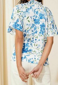 Žena v bílé blůze s modrým květinovým potiskem a volnými rukávy, spojená s krémovými kalhotami, stojí s rukama za zády.