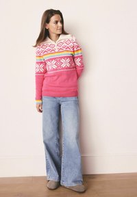 Roze gebreide trui met witte geometrische patronen en regenboogstrepen, gecombineerd met lichtblauwe flared jeans en grijze enkellaarsjes.