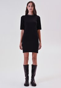 Vestito nero a maniche corte con una forma aderente, caratterizzato da un collo con gioielli. Modella con stivali neri alti. Texture del tessuto liscia.