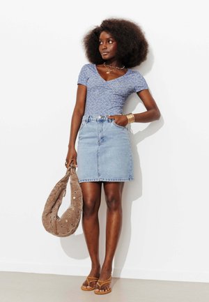 Femme avec coiffure afro porte un haut bleu à motifs, une jupe en jean, des sandales, des bijoux en or, et tient un sac à main en daim marron contre un mur blanc.