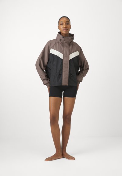 Braune und schwarze wasserdichte Jacke mit weißen Streifendetails, verstellbare Kapuze und elastische Bündchen. Das Model trägt schwarze Shorts.