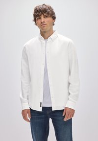 Weißes Langarmhemd mit Knopfleiste, Kragen und texturiertem Stoff, getragen über einem weißen T-Shirt und kombiniert mit blauen Jeans.