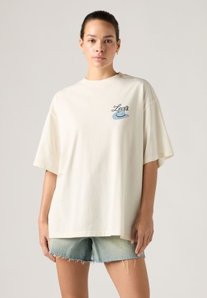 Femme portant un t-shirt blanc ample Levi's avec un logo de chapeau bleu et un short en jean clair, debout devant un fond uni.