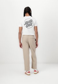 Camiseta de algodão branca com um logótipo circular preto nas costas, combinada com calças de alfaiataria bege e ténis com riscas vermelhas.