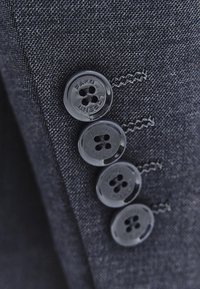 Tkanina garniturowa w ciemnoszarym kolorze o teksturowanym splocie, z czterema czarnymi guzikami przyszytymi do mankietu. Guziki mają czterodziurkowy wzór i grawerowaną etykietę.